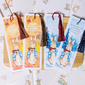 May include: Four illustrated bookmarks with a rabbit in a blue jacket and red scarf. Each bookmark has a different background and tassel colour, with text: "The coziest burrow is lined with books and blankets" and "Snowflakes and storybooks make the best kind of day."