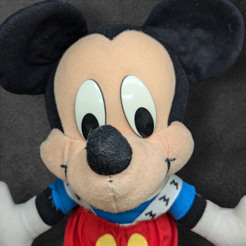 Pu&ograve; includere: Un giocattolo di peluche di Topolino con orecchie nere, grandi occhi bianchi e un naso nero. Indossa una camicia a righe blu e bianche con una fascia rossa e gialla su cui &egrave; scritto "MM".