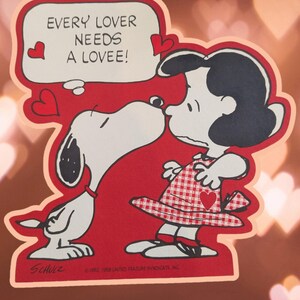 May include: A red and white Valentine's Day card featuring Snoopy and his girlfriend, Lucy. The card has a speech bubble that says "Every lover needs a lovee!" with hearts around it.