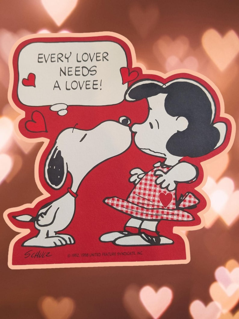 May include: A red and white Valentine's Day card featuring Snoopy and his girlfriend, Lucy. The card has a speech bubble that says "Every lover needs a lovee!" with hearts around it.