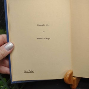 May include: Open book with text reading "Copyright, 1939 by Pundit Acharya" and "First Print." The book has a blue cover and is held open by a wooden book stand. The pages are a light tan color.
