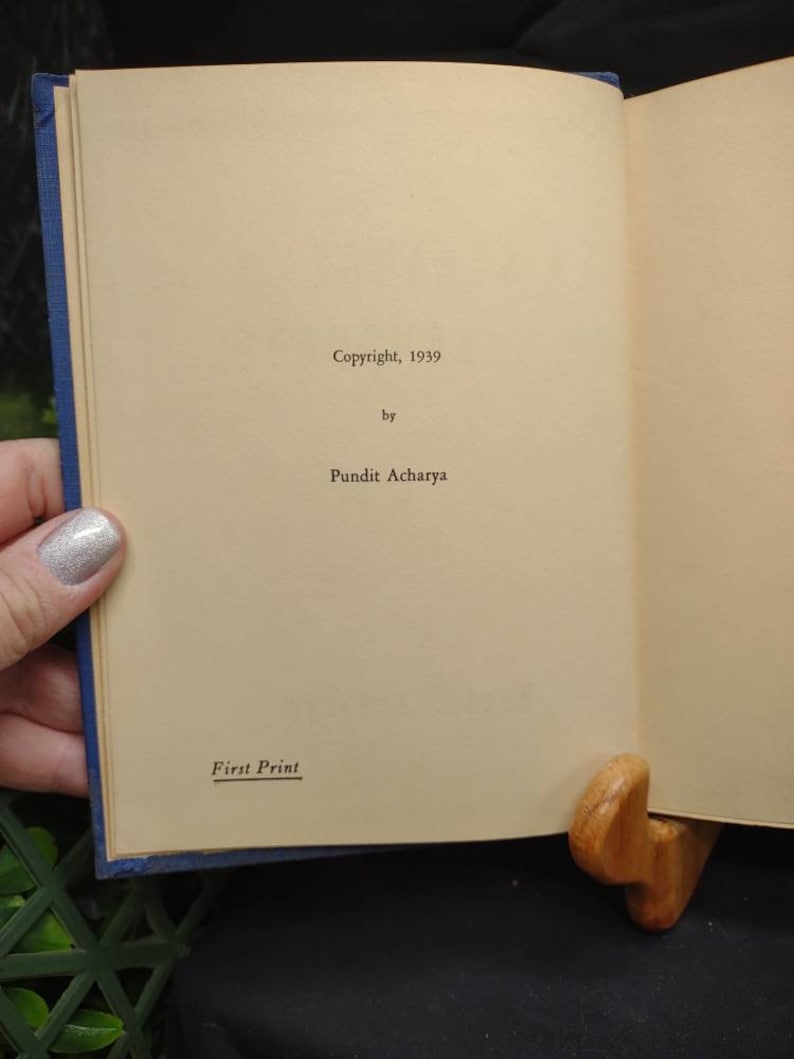 May include: Open book with text reading "Copyright, 1939 by Pundit Acharya" and "First Print." The book has a blue cover and is held open by a wooden book stand. The pages are a light tan color.