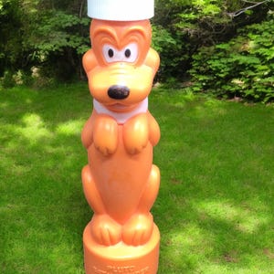 May include: Orange Pluto bath powder bottle with a white lid. The bottle is shaped like Pluto, a cartoon dog character. The base of the bottle has the text "Pluto Bath Powder". The bottle is standing on green grass.