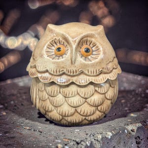 May include: A ceramic owl figurine with a tan glaze. The owl has large, round eyes with orange irises and black pupils. The body is detailed with a scalloped design. The owl is a decorative item.
