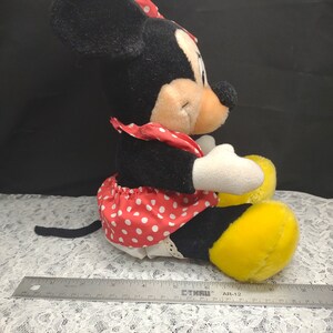 Op de afbeelding: Een pluche zwart-witte Minnie Mouse speelgoed dat een rode jurk draagt met witte stippen en een rode strik. Het speelgoed zit met gekruiste benen en heeft gele schoenen.