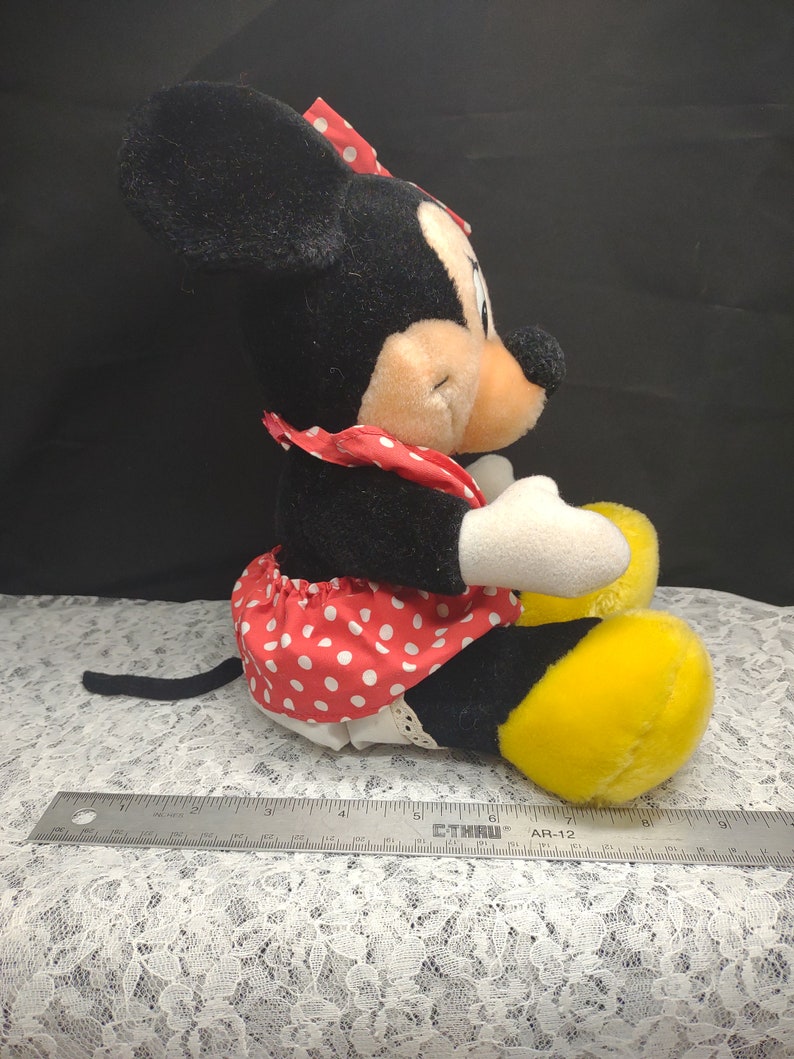 Op de afbeelding: Een pluche zwart-witte Minnie Mouse speelgoed dat een rode jurk draagt met witte stippen en een rode strik. Het speelgoed zit met gekruiste benen en heeft gele schoenen.