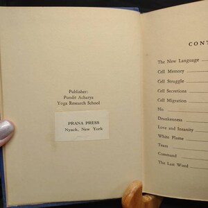 May include: Open book with text, held open by a wooden book stand. The left page displays the publisher's information: PRANA PRESS, Nyack, New York. The right page lists the table of contents, including "Cell Memory" and "White Flame."