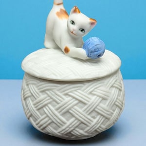 May include: A white ceramic trinket box with a woven basket design. A white and orange cat figurine is perched on top, playing with a blue ball of yarn. The cat has green eyes and is in a playful pose.