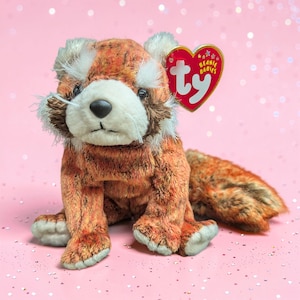 May include: A plush red panda toy with textured orange, brown, and white fur, featuring a white muzzle, black nose, and dark eyes. A heart-shaped red tag with the white text ty and Beanie Babies is attached to the ear of the stuffed animal.