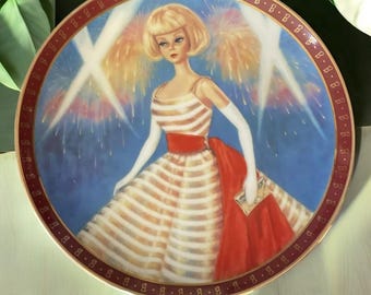 May include: A porcelain plate featuring a blonde doll wearing a white and red striped dress with a red sash. The doll is standing in front of a blue background with fireworks and spotlights. The plate has a gold rim with a repeating pattern of "B"s.