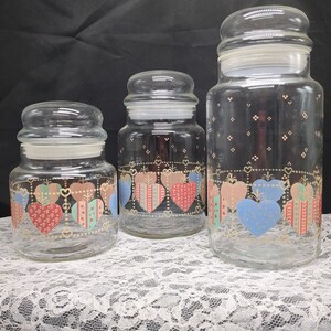May include: Set of three glass canisters with heart-shaped designs in pink, blue, and green. The canisters have gold accents and are arranged in order of size from smallest to largest.