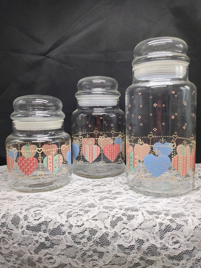 May include: Set of three glass canisters with heart-shaped designs in pink, blue, and green. The canisters have gold accents and are arranged in order of size from smallest to largest.