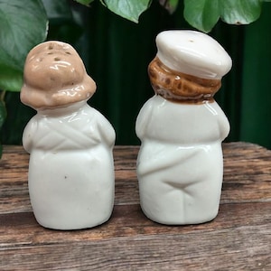 May include: A pair of ceramic salt and pepper shakers shaped like people wearing hats. The salt shaker is a woman with a brown hat and the pepper shaker is a man with a white hat. Both shakers are white with brown accents.