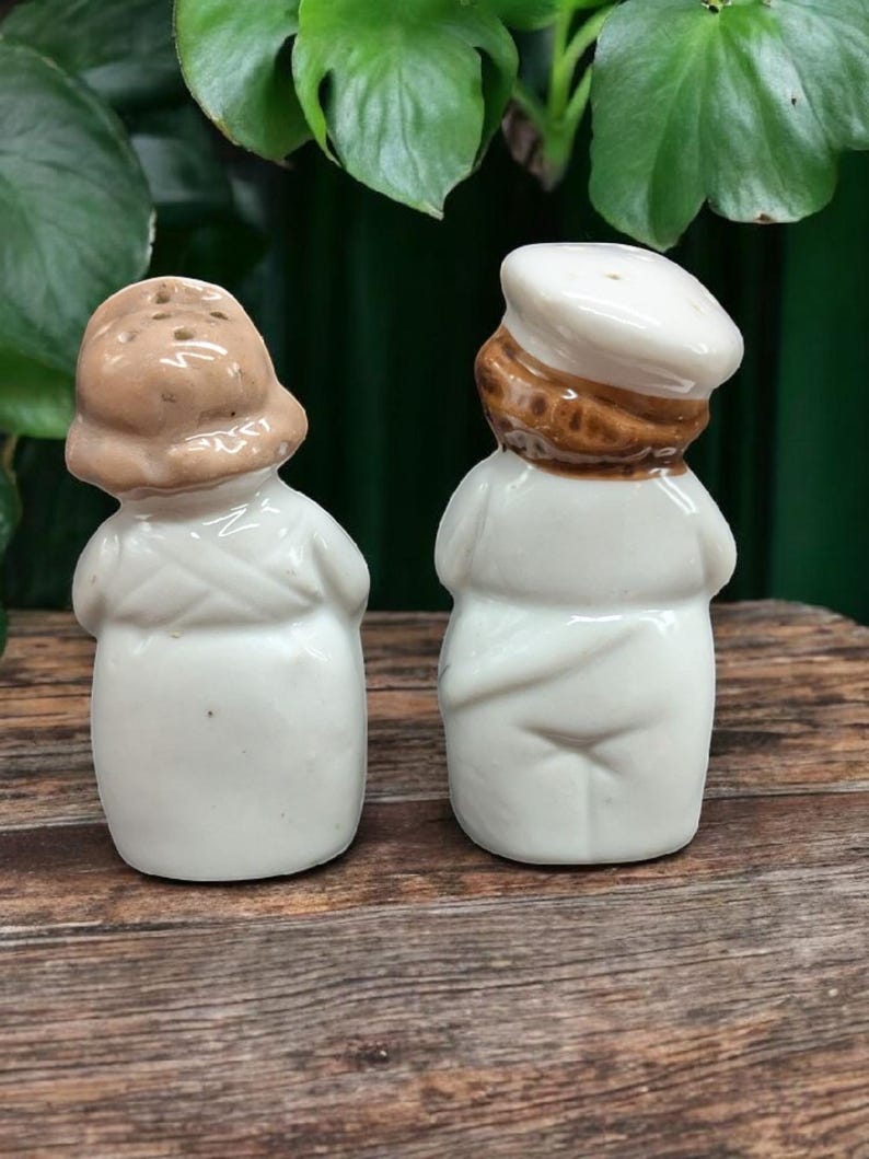 May include: A pair of ceramic salt and pepper shakers shaped like people wearing hats. The salt shaker is a woman with a brown hat and the pepper shaker is a man with a white hat. Both shakers are white with brown accents.