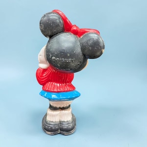 Op de afbeelding: Vintage Minnie Mouse beeldje. Het beeldje heeft zwarte oren, een rode strik, een rode jurk, een blauwe rok en zwarte schoenen. Het plastic beeldje heeft het Disney-logo op de achterkant.