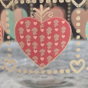May include: A clear glass jar with a gold heart design. The heart is filled with a red and gold pattern. The jar has a gold rim and a gold dot pattern around the top and bottom.