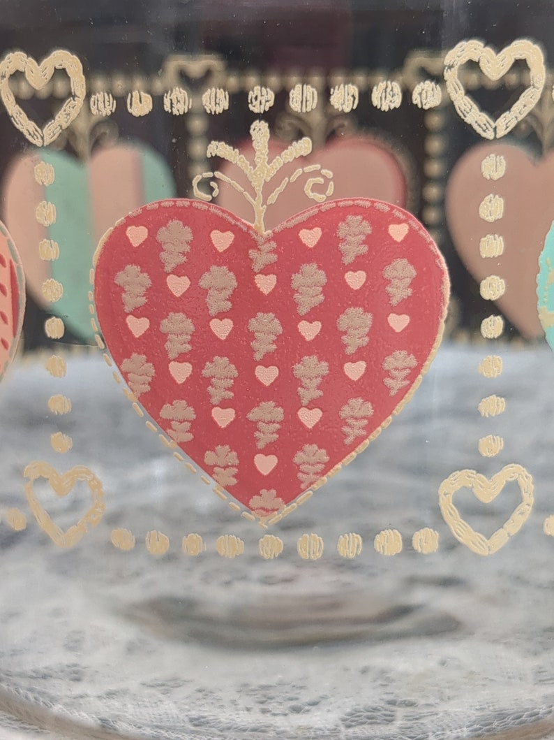 May include: A clear glass jar with a gold heart design. The heart is filled with a red and gold pattern. The jar has a gold rim and a gold dot pattern around the top and bottom.