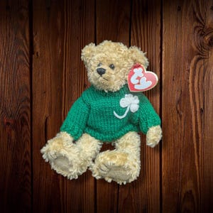 May include: A plush teddy bear with tan fur and a green sweater featuring a white shamrock. The bear has a black nose and eyes, and a red heart-shaped tag. The background is a dark wood grain.