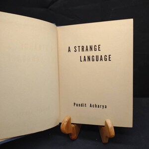 May include: An open book with the title "A Strange Language" in black text on a cream-colored page. The author's name, Pundit Acharya, is below the title. The book is propped up by a wooden book stand.