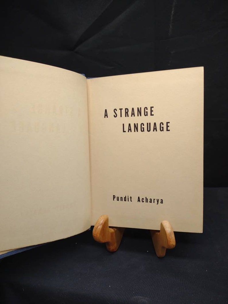 May include: An open book with the title "A Strange Language" in black text on a cream-colored page. The author's name, Pundit Acharya, is below the title. The book is propped up by a wooden book stand.
