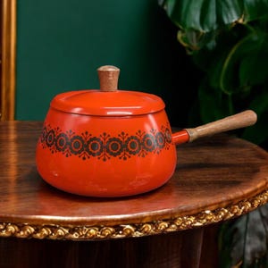 May include: A vintage red enamel saucepan with a wooden handle and lid. The pot features a black floral pattern around the base. The lid has a wooden knob. The saucepan is sitting on a wooden table.