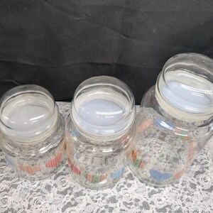 May include: Three glass jars with lids, featuring a colorful pattern of houses and hearts. The jars are clear glass and the lids are white.