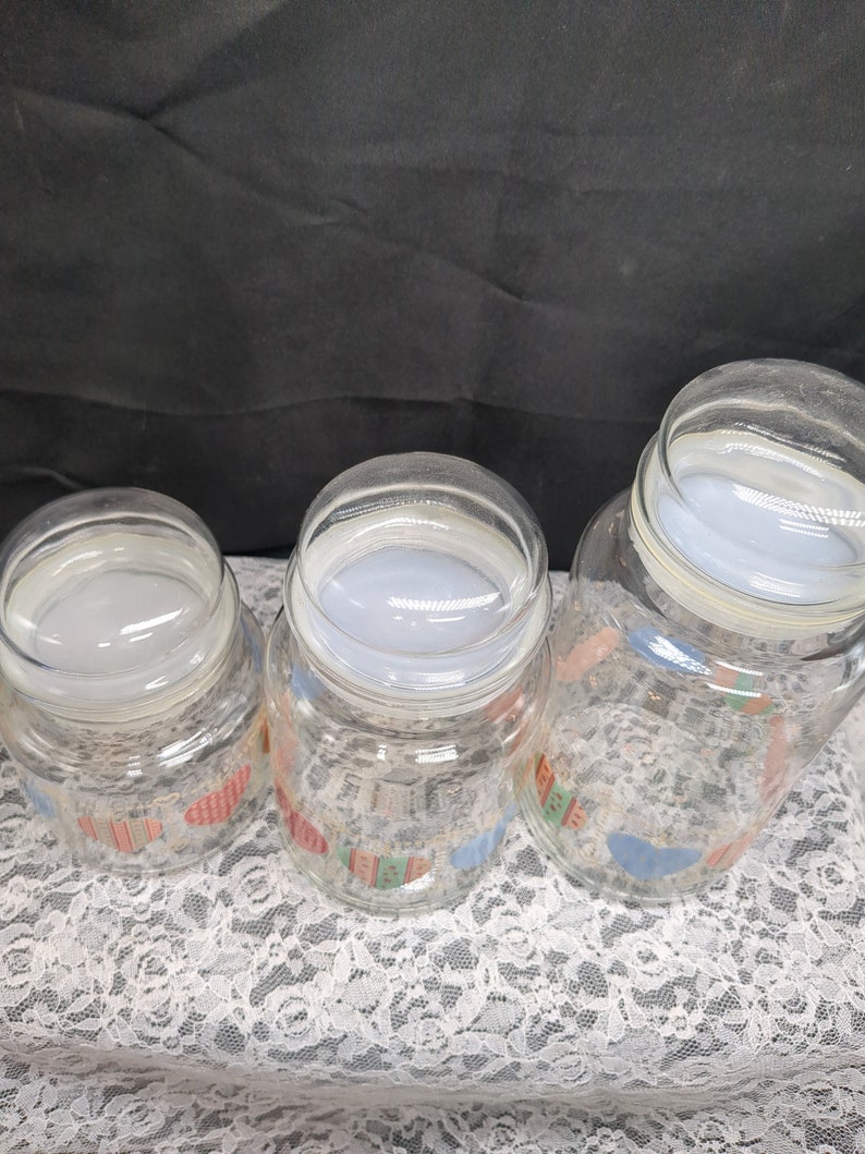 May include: Three glass jars with lids, featuring a colorful pattern of houses and hearts. The jars are clear glass and the lids are white.