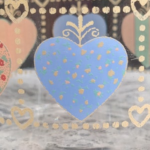 May include: A glass jar with a blue heart design. The heart is decorated with a floral pattern in yellow, pink, and green. The jar has a gold border with a repeating pattern of hearts and dots.