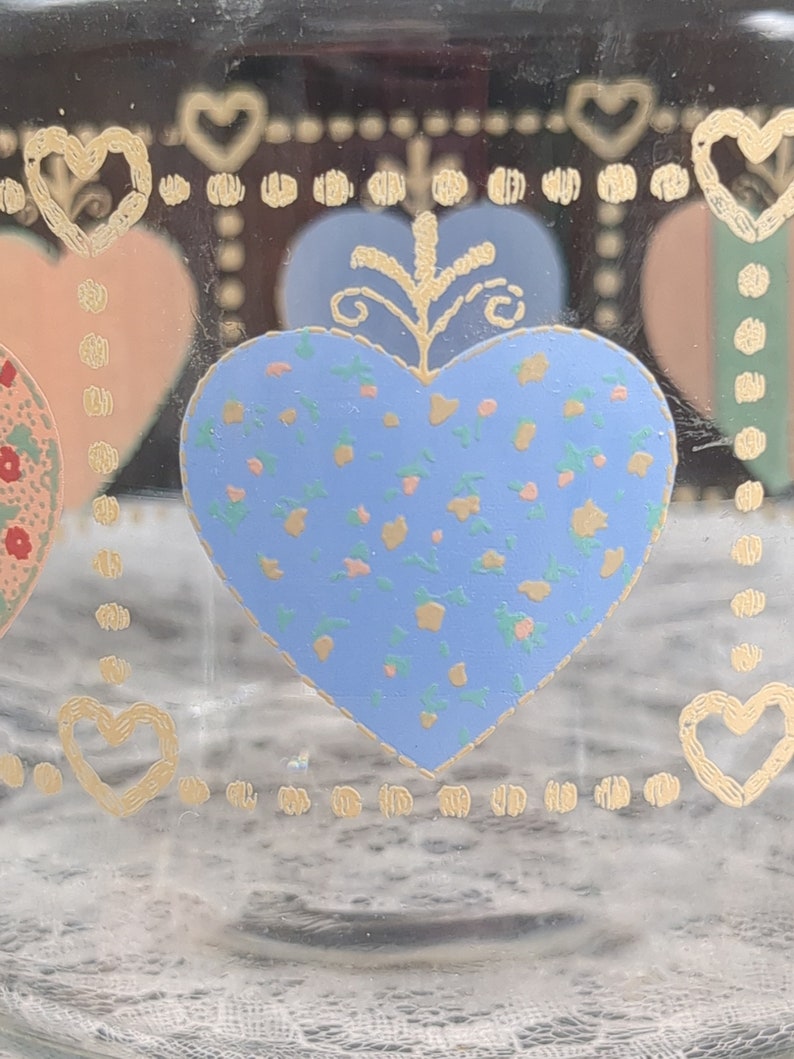 May include: A glass jar with a blue heart design. The heart is decorated with a floral pattern in yellow, pink, and green. The jar has a gold border with a repeating pattern of hearts and dots.