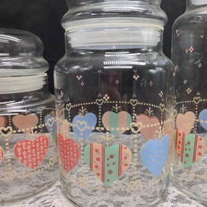 May include: Set of three glass canisters with heart-shaped designs. The canisters are clear glass with a white lid and feature a colorful patchwork heart design with pink, red, blue, and green accents.