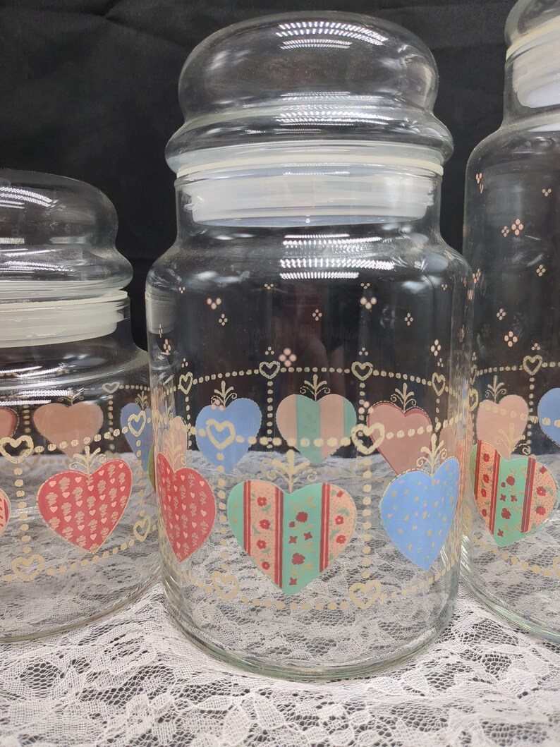 May include: Set of three glass canisters with heart-shaped designs. The canisters are clear glass with a white lid and feature a colorful patchwork heart design with pink, red, blue, and green accents.