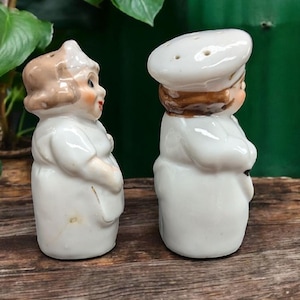 May include: A set of two white ceramic salt and pepper shakers shaped like people. The woman is wearing a white dress and a brown hat. The man is wearing a white chef's hat and a white uniform.