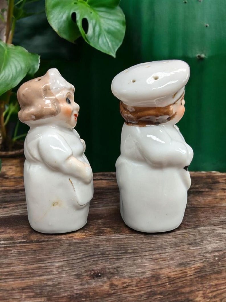 May include: A set of two white ceramic salt and pepper shakers shaped like people. The woman is wearing a white dress and a brown hat. The man is wearing a white chef's hat and a white uniform.
