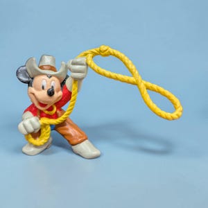 Op de afbeelding: Een Mickey Mouse-beeldje verkleed als cowboy, met een hoed, rood shirt, bruine broek en witte laarzen. Het figuur houdt een gele lasso vast tegen een lichtblauwe achtergrond.