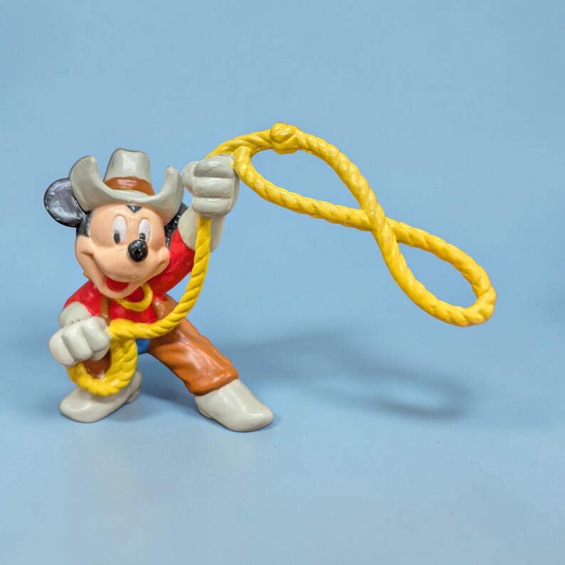 Op de afbeelding: Een Mickey Mouse-beeldje verkleed als cowboy, met een hoed, rood shirt, bruine broek en witte laarzen. Het figuur houdt een gele lasso vast tegen een lichtblauwe achtergrond.