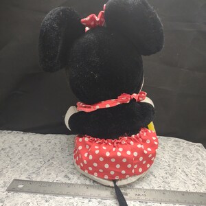 Op de afbeelding: Een zwart en rood pluche speelgoed van een cartoonmuis die een rode jurk met witte stippen en een rode strik draagt. Het speelgoed heeft een lange zwarte staart.