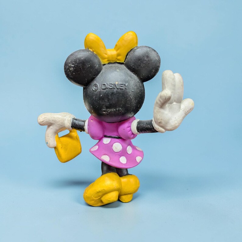 Op de afbeelding: Een Minnie Mouse-beeldje met een gele strik, gele schoenen en een roze jurk met witte stippen. Het beeldje houdt een gele handtas vast en heeft witte handschoenen. Het Disney-logo is zichtbaar op de achterkant.