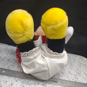 Op de afbeelding: Een pluche speelgoed van een cartoonfiguur dat een rood-witte jurk met zwarte stippen draagt. Het speelgoed heeft gele oren en zwarte schoenen. Het speelgoed zit op een witte kanten stof. Het label van het speelgoed luidt "Disney's Mickey Mouse Clubhouse, 2012, Gemaakt in China, Allemaal nieuw materiaal, Reg. Nr. PA-11497 (CN)"