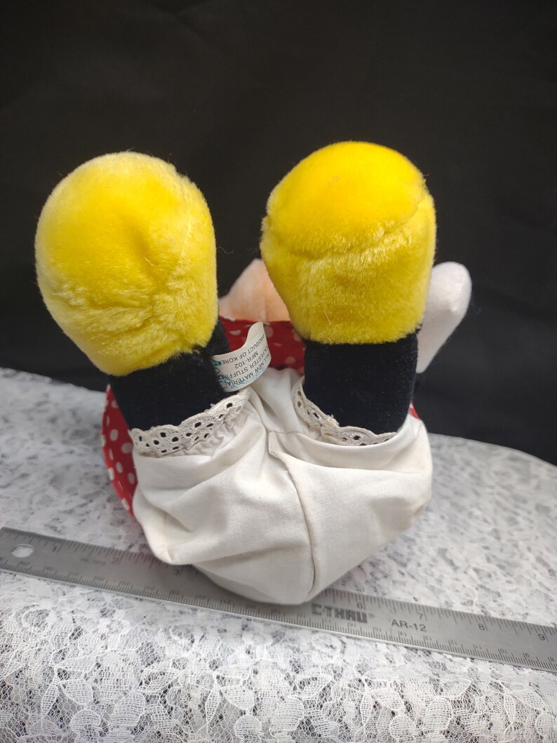 Op de afbeelding: Een pluche speelgoed van een cartoonfiguur dat een rood-witte jurk met zwarte stippen draagt. Het speelgoed heeft gele oren en zwarte schoenen. Het speelgoed zit op een witte kanten stof. Het label van het speelgoed luidt "Disney's Mickey Mouse Clubhouse, 2012, Gemaakt in China, Allemaal nieuw materiaal, Reg. Nr. PA-11497 (CN)"