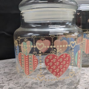 May include: A set of three clear glass canisters with heart-shaped designs in pink, blue, green, and red. The canisters have gold accents and a white background.