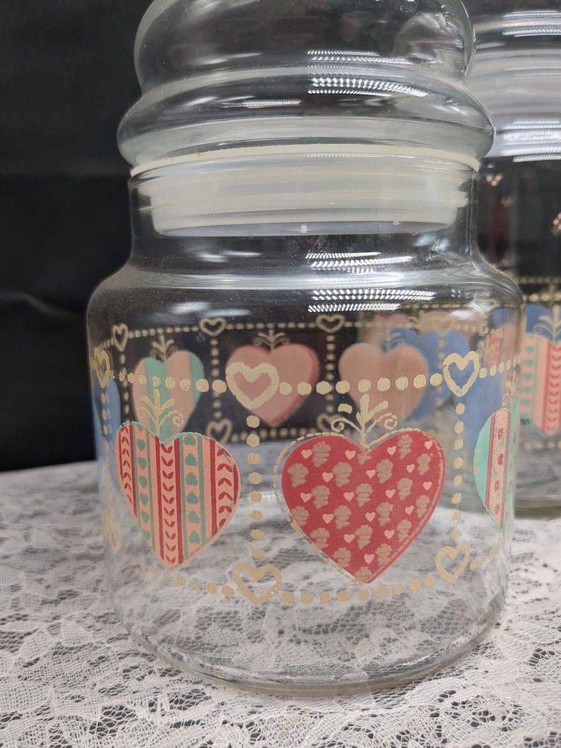 May include: A set of three clear glass canisters with heart-shaped designs in pink, blue, green, and red. The canisters have gold accents and a white background.