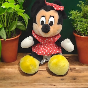 Op de afbeelding: Een pluche speelgoed van Minnie Mouse met een rode jurk met witte stippen en een rode strik. Het speelgoed heeft gele schoenen en zwarte ogen.