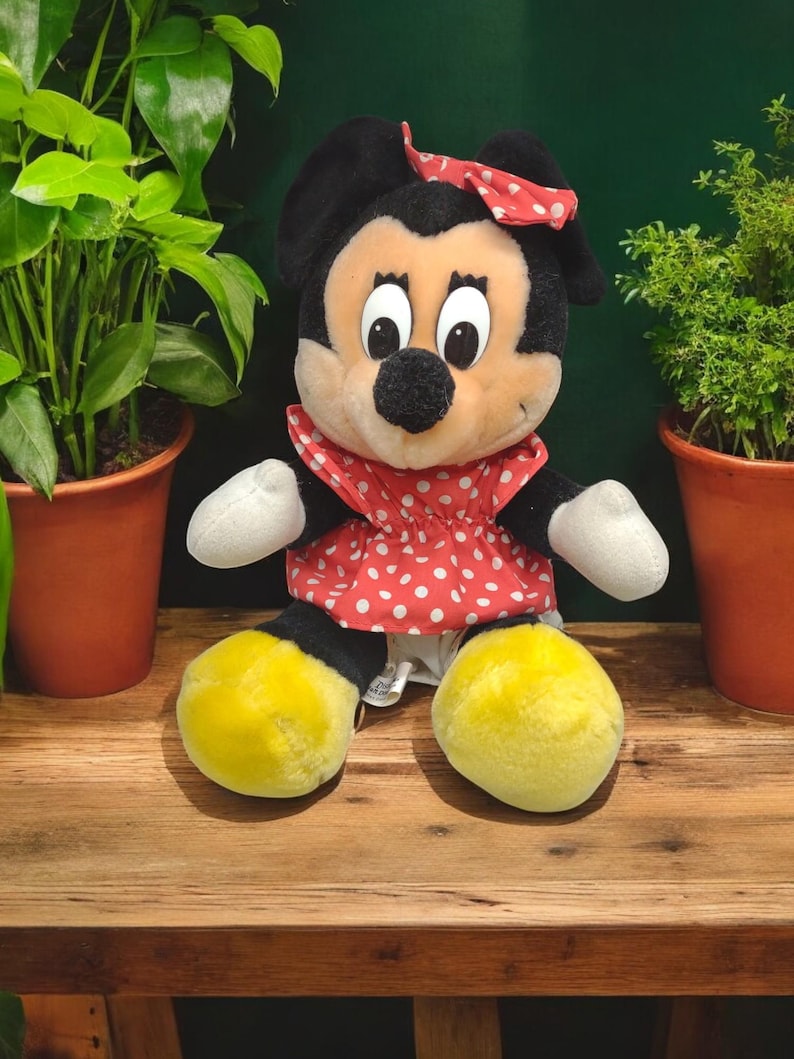 Op de afbeelding: Een pluche speelgoed van Minnie Mouse met een rode jurk met witte stippen en een rode strik. Het speelgoed heeft gele schoenen en zwarte ogen.