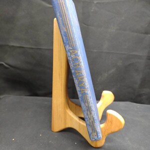 May include: A blue book titled "ACHARYA" with gold lettering, displayed on a wooden book stand. The book has a textured cover and is positioned at an angle, showcasing its spine. The stand is made of light-colored wood.