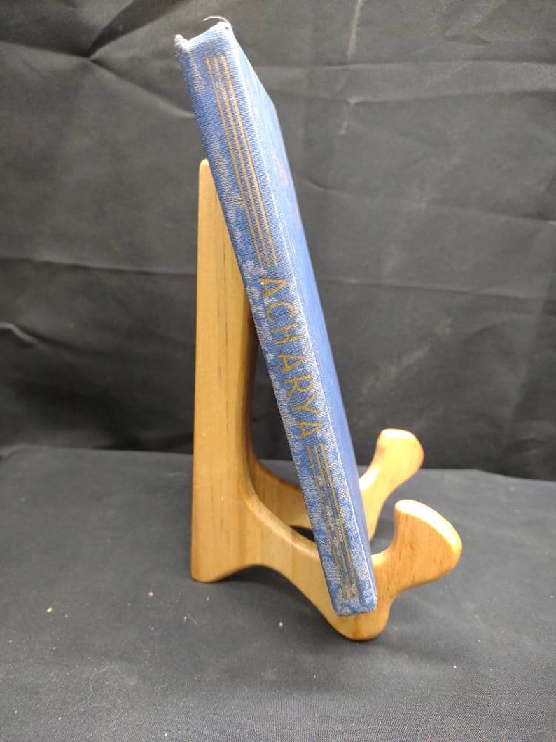 May include: A blue book titled "ACHARYA" with gold lettering, displayed on a wooden book stand. The book has a textured cover and is positioned at an angle, showcasing its spine. The stand is made of light-colored wood.