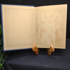 May include: An open book with aged, cream-colored pages and a blue cover. The book is propped open on a wooden stand, revealing the interior pages. The pages show signs of age, with slight discoloration. The book is set against a dark background.