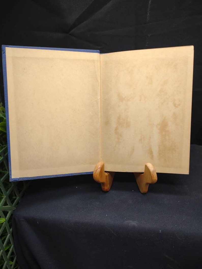 May include: An open book with aged, cream-colored pages and a blue cover. The book is propped open on a wooden stand, revealing the interior pages. The pages show signs of age, with slight discoloration. The book is set against a dark background.