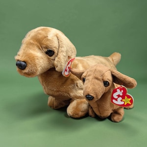 May include: Two plush dachshund dog toys in shades of brown. The larger dog is light brown, and the smaller one is a darker brown. Both have black eyes and noses, and red heart-shaped tags.