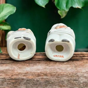 May include: A pair of vintage ceramic salt and pepper shakers shaped like people. The shakers are white with brown accents and have a hand-painted design. The bottom of each shaker is marked "JAPAN".