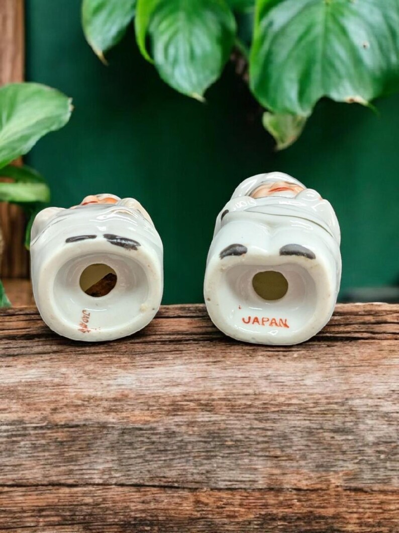 May include: A pair of vintage ceramic salt and pepper shakers shaped like people. The shakers are white with brown accents and have a hand-painted design. The bottom of each shaker is marked "JAPAN".